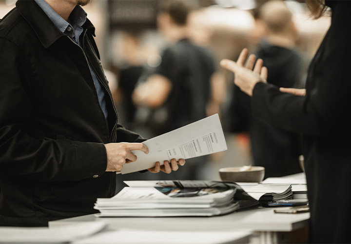 Woman talking to man at tradeshow after offering him a company brochure.